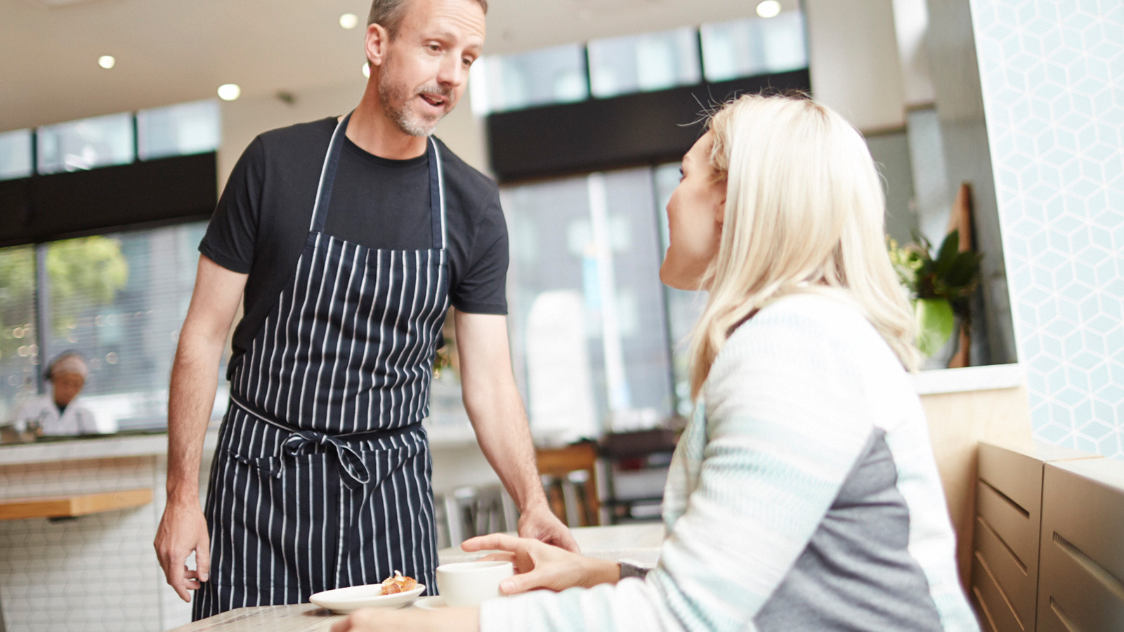 A man talking to a woman seated at a table in a café.